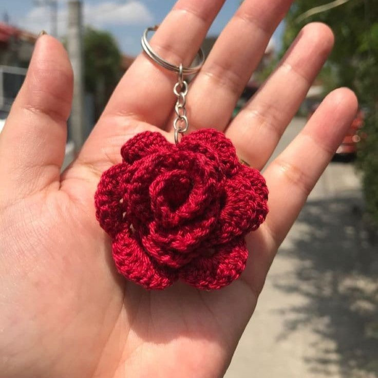 Red crocheted rose keychain held in a hand with a blurred outdoor background