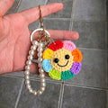 Hand holding a keychain with a colorful smiley face charm and pearl bracelet on a tiled floor background.