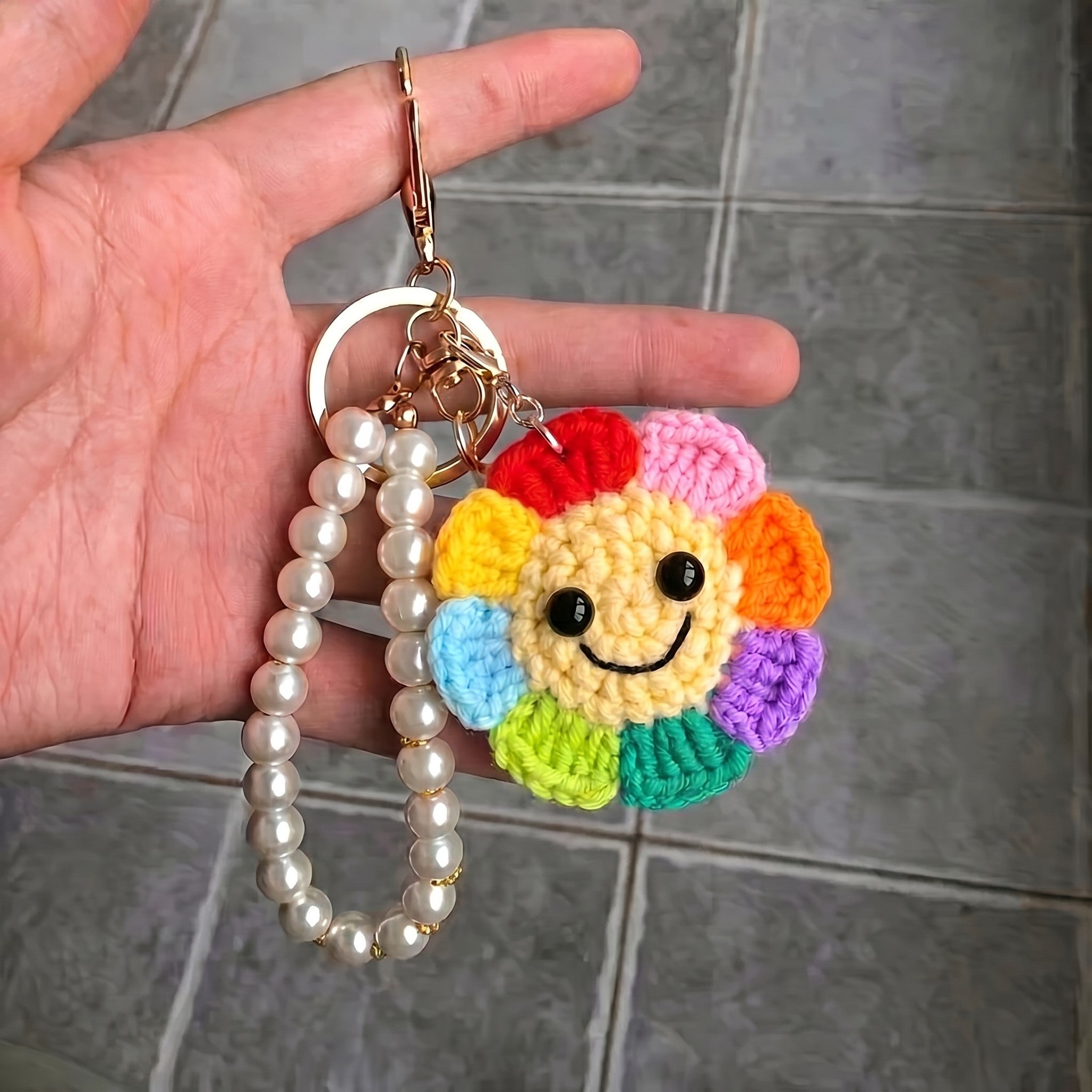 Hand holding a keychain with a colorful smiley face charm and pearl bracelet on a tiled floor background.